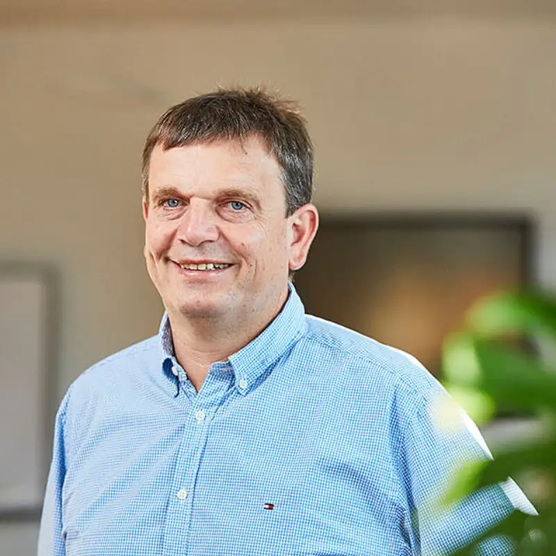 Ein Portrait von Stefan Suhr, von Endress Hauser, der lächelnd in einem sonnigen Büro hinter einer grünen Pflanze steht Ein Portrait von Stefan Suhr, von Endress Hauser, der lächelnd in einem sonnigen Büro hinter einer grünen Pflanze steht